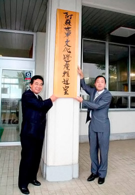 阿蘇世界文化遺産推進室の看板の前にたつ佐藤阿蘇市長と阿南熊本県教育次長